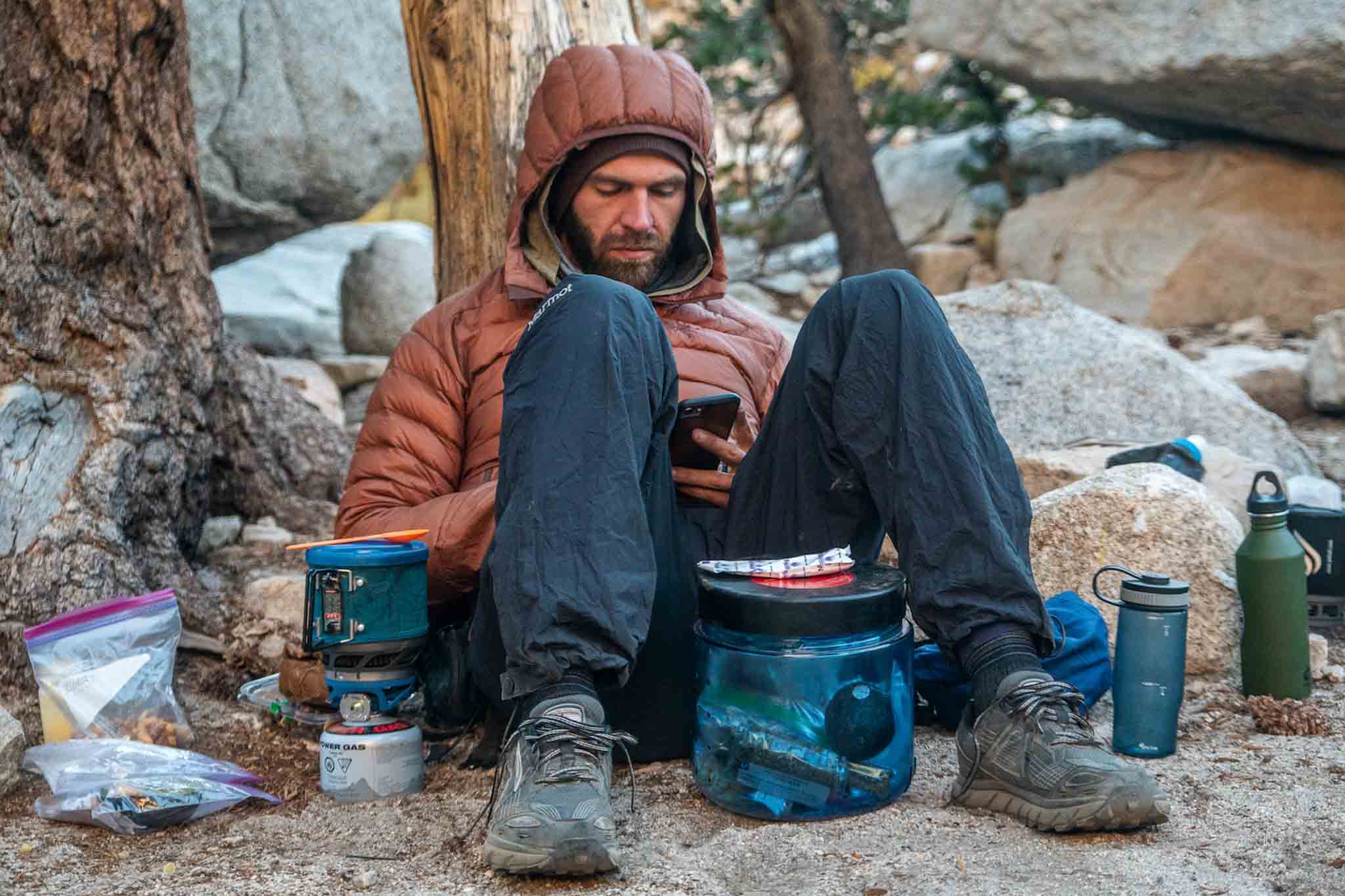 Man sitting against a tree, BearVault bear canister at his feet, stove next to him.
