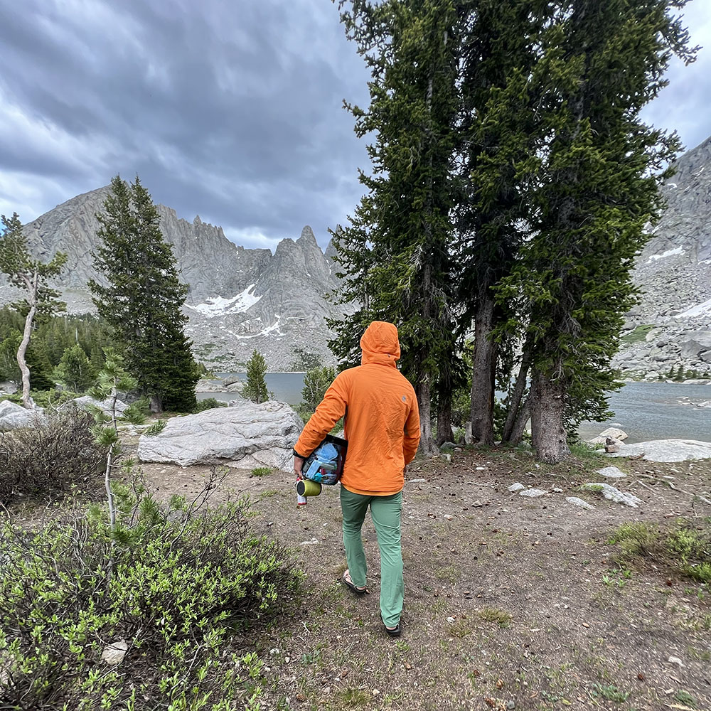 A man walking toward a lake surrounded by mountains with a bear canister under his arm