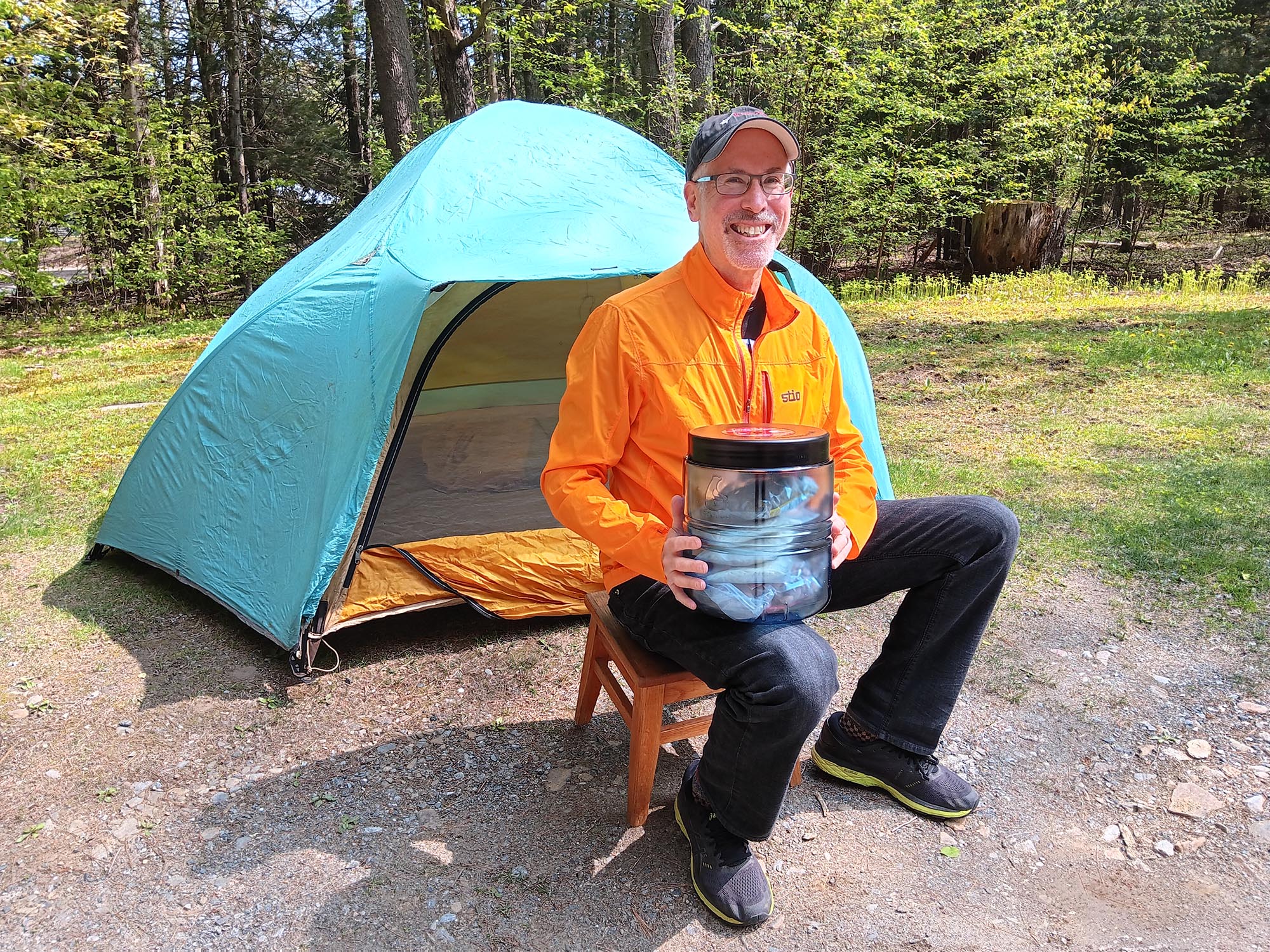 A man sits on a stool in front of a tent with his bear proof container